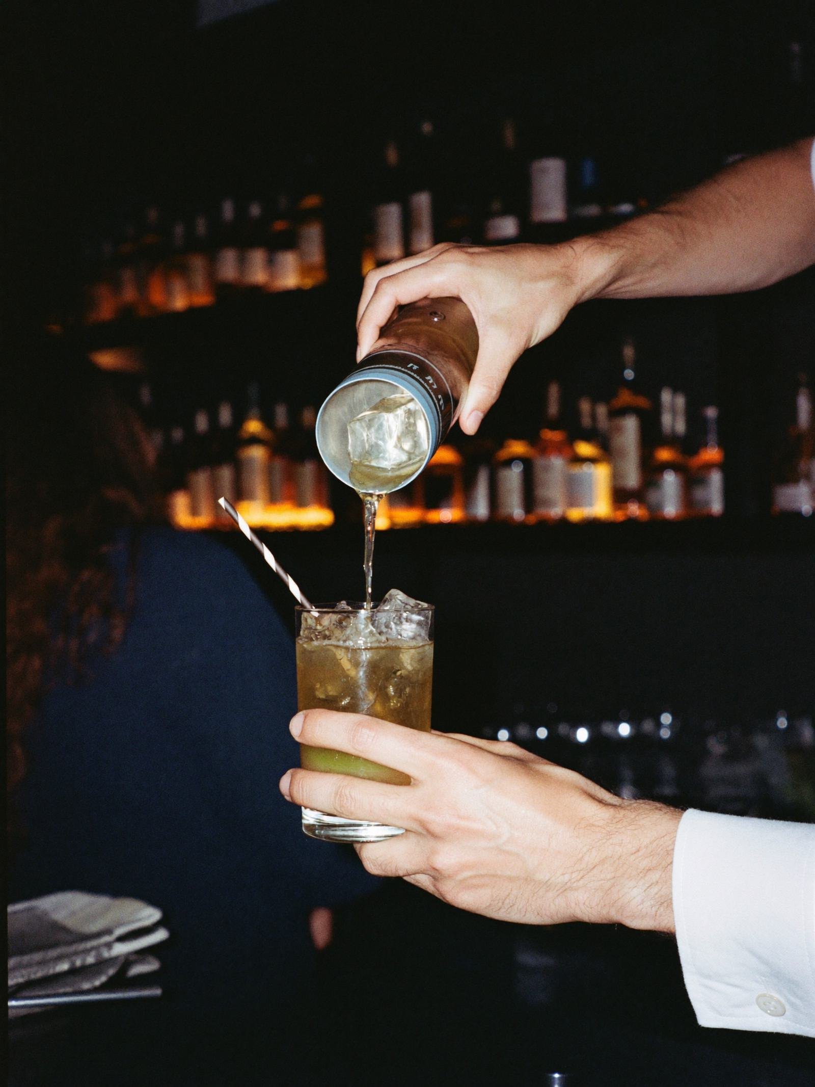 Les mains d'un barman filtrent un cocktail devant un mur de bouteilles.