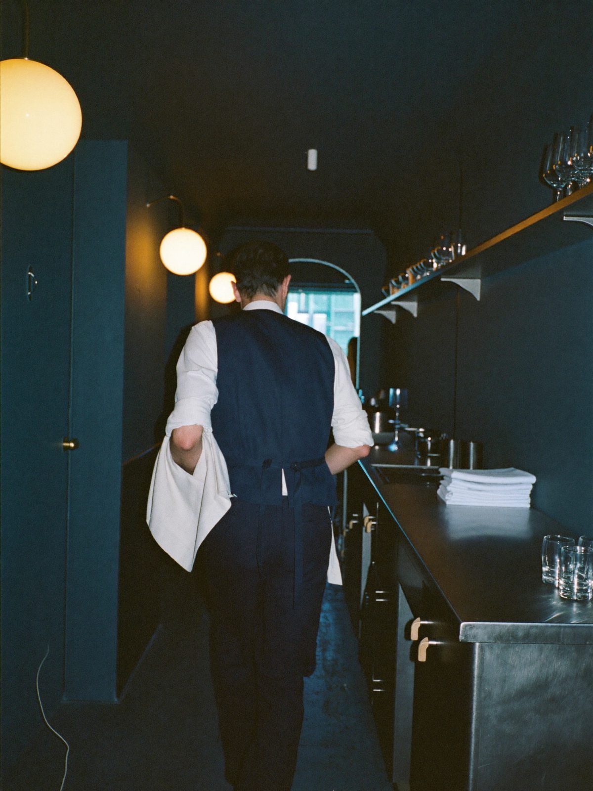 Un serveur Flexho, une pile de linge à la main, remonte le couloir de service avant l'ouverture.