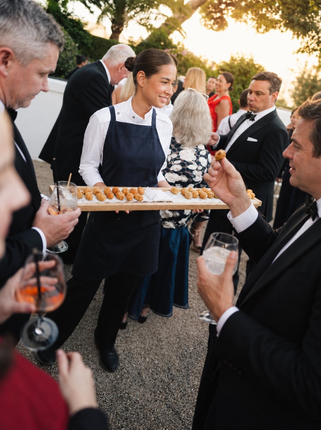 Une serveuse Flexho, en tablier bleu marine, présente un plateau de croquettes aux invités d'une réception en extérieur.